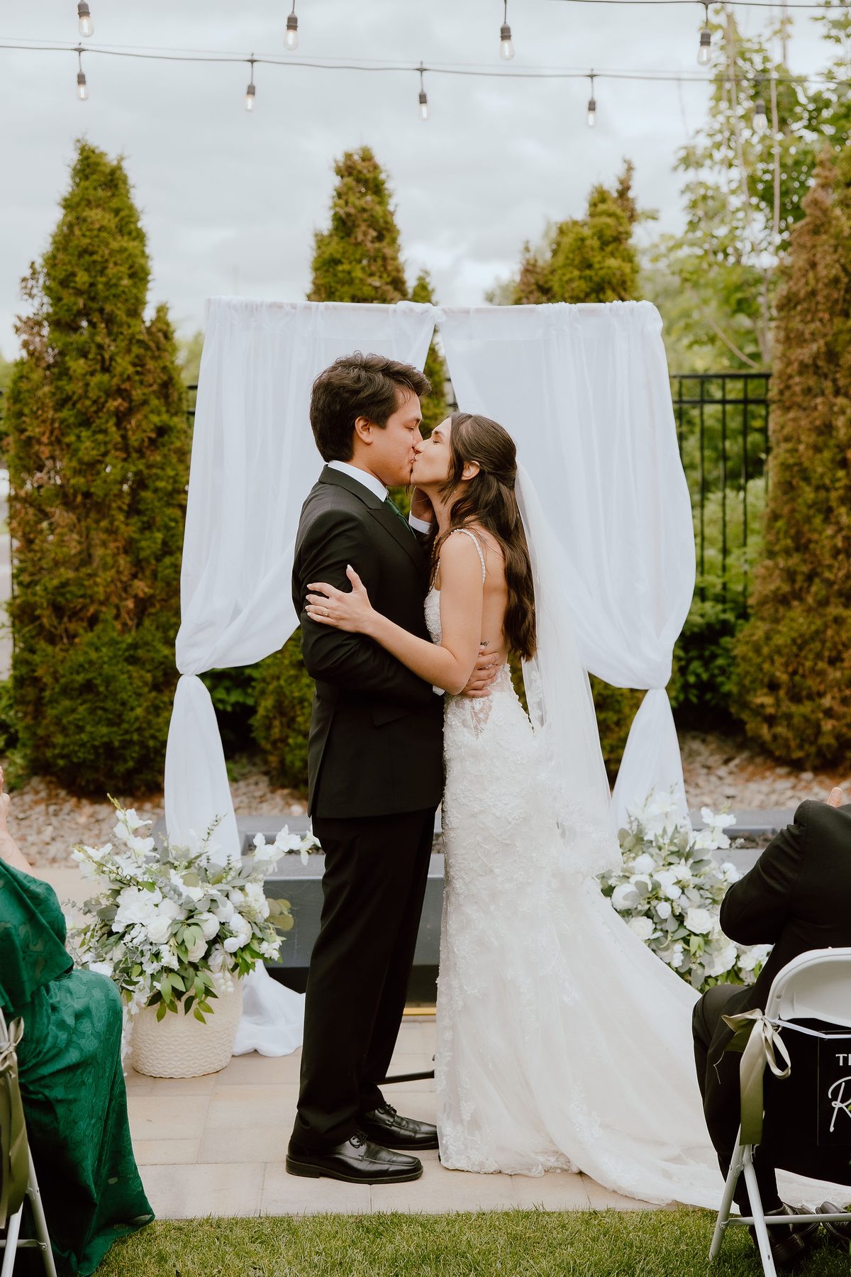 An outdoor ceremony under a white draped arch of roses and eucalyptus and string lights, the couple kissing as the bride's fitted lace gown and long veil trail behind her