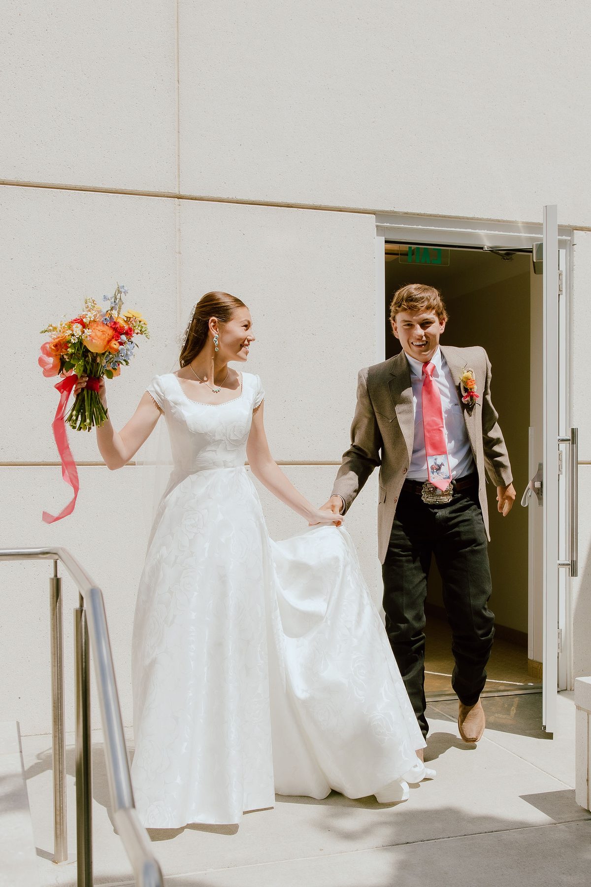 A smiling bride and groom walking hand in hand out through a doorway, the bride holding up the train of her lace gown and carrying a bright bouquet