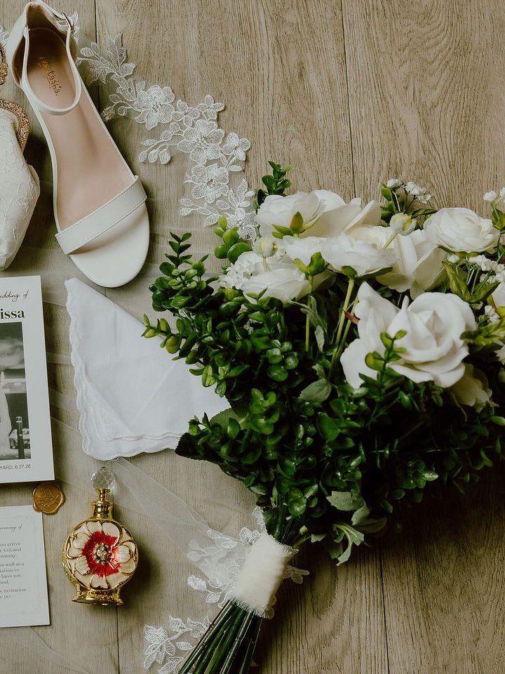 A close vignette of a white rose and eucalyptus bouquet beside a single bridal heel, lace veil trim and a gold perfume bottle