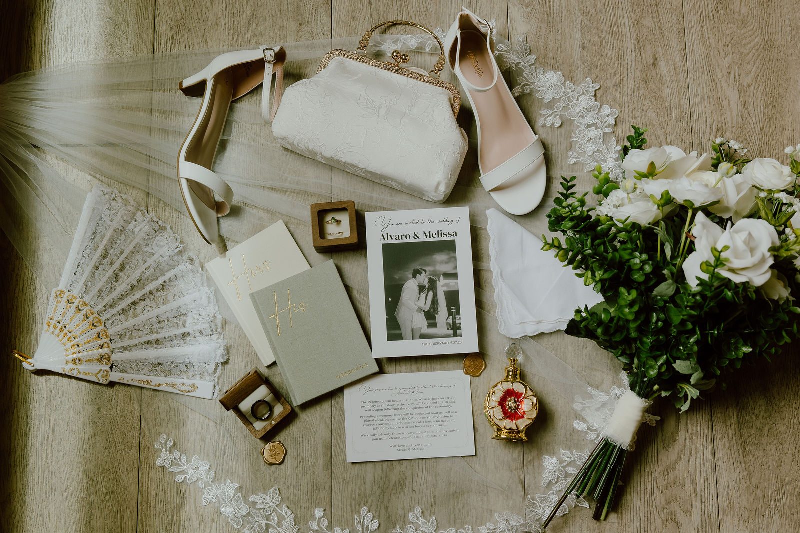 A wide overhead flat lay on a warm wood floor with bridal heels, a beaded clutch, a veil, an invitation suite, a vow book, rings, a lace fan, a gold locket and a white rose bouquet