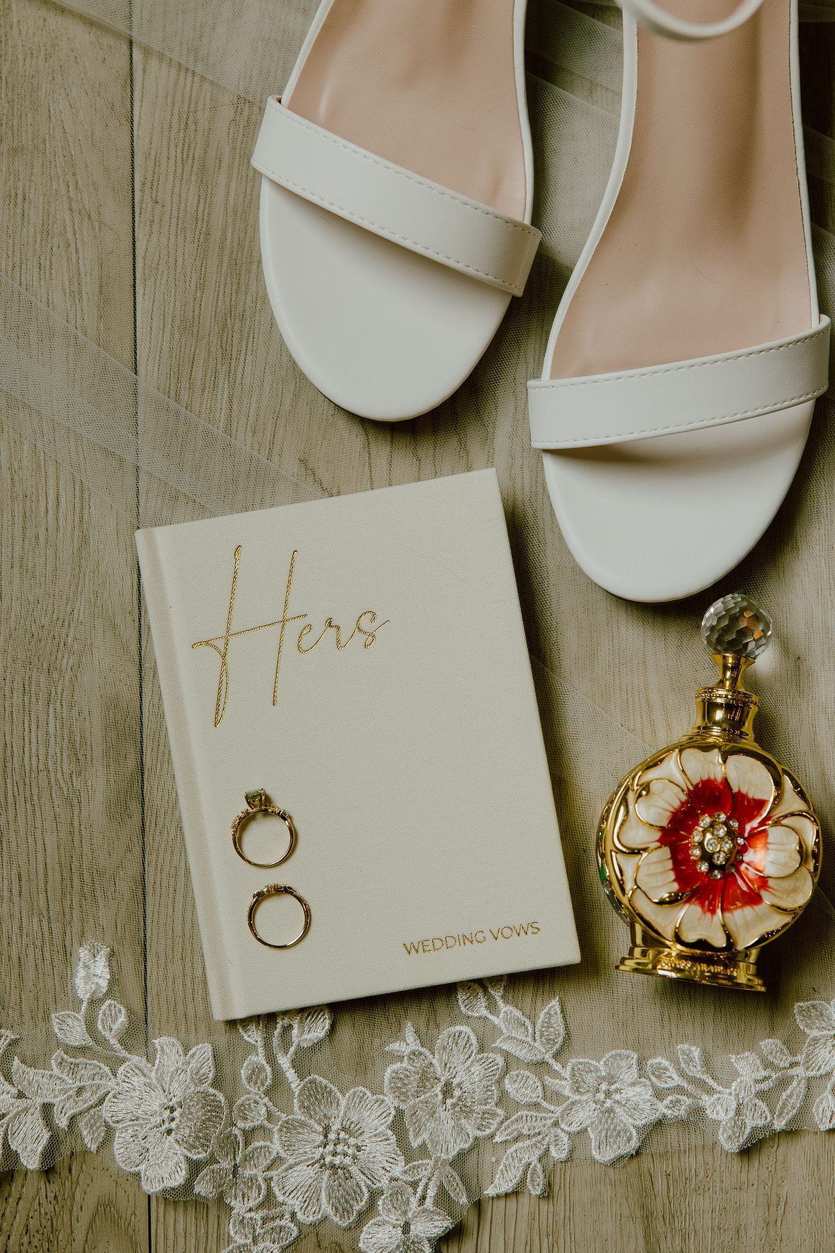 A close detail of white bridal heels, a vow book, wedding rings and a gold locket resting on lace over concrete