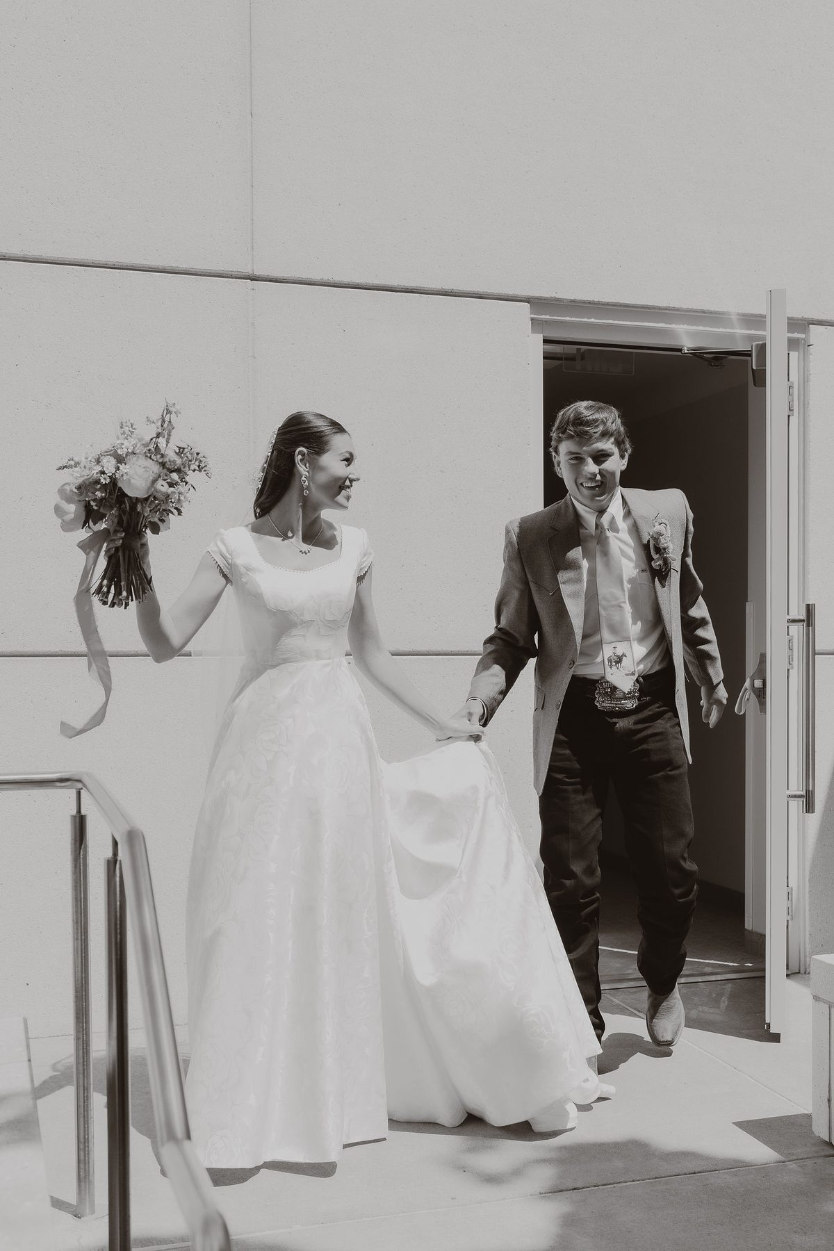 A black-and-white frame of a bride and groom stepping out through a doorway, the lace of her gown and its train catching the light