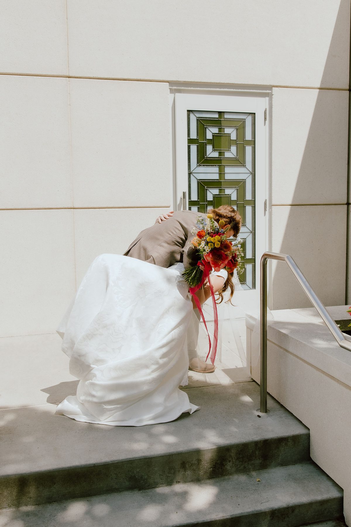 A bride in a full brocade gown leaning toward her groom in a doorway, her skirt spread across concrete steps, holding a bright red and orange bouquet